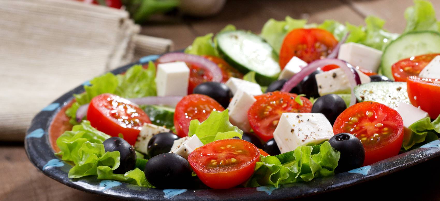 Uma imagem em close-up de uma salada grega fresca. A salada está em um prato azul-escuro com bordas decoradas, em um fundo de madeira. Ela contém alface verde, rodelas de pepino, azeitonas pretas, cubos de queijo feta e tomates-cereja cortados ao meio. A luz ilumina a salada, destacando a textura do queijo e o brilho dos tomates.